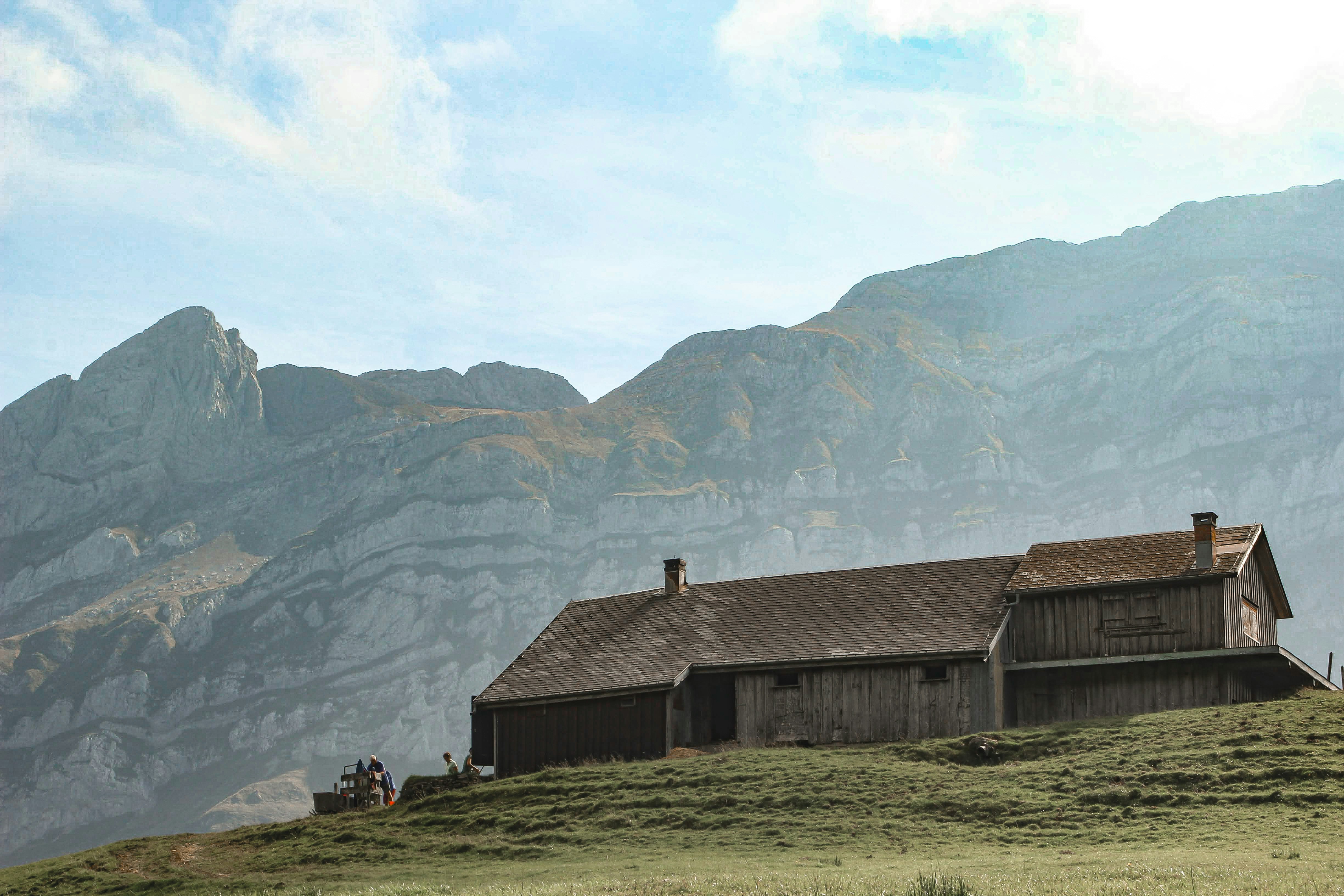 Wooden cabin on a grassy hill with mountains behind.