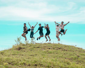 Six friends jumping joyfully on a grassy hill.