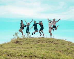 Six friends jumping joyfully on a grassy hill.