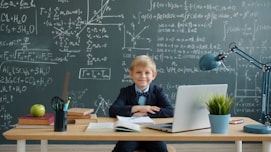 Young boy smiles at desk with laptop and chalkboard.