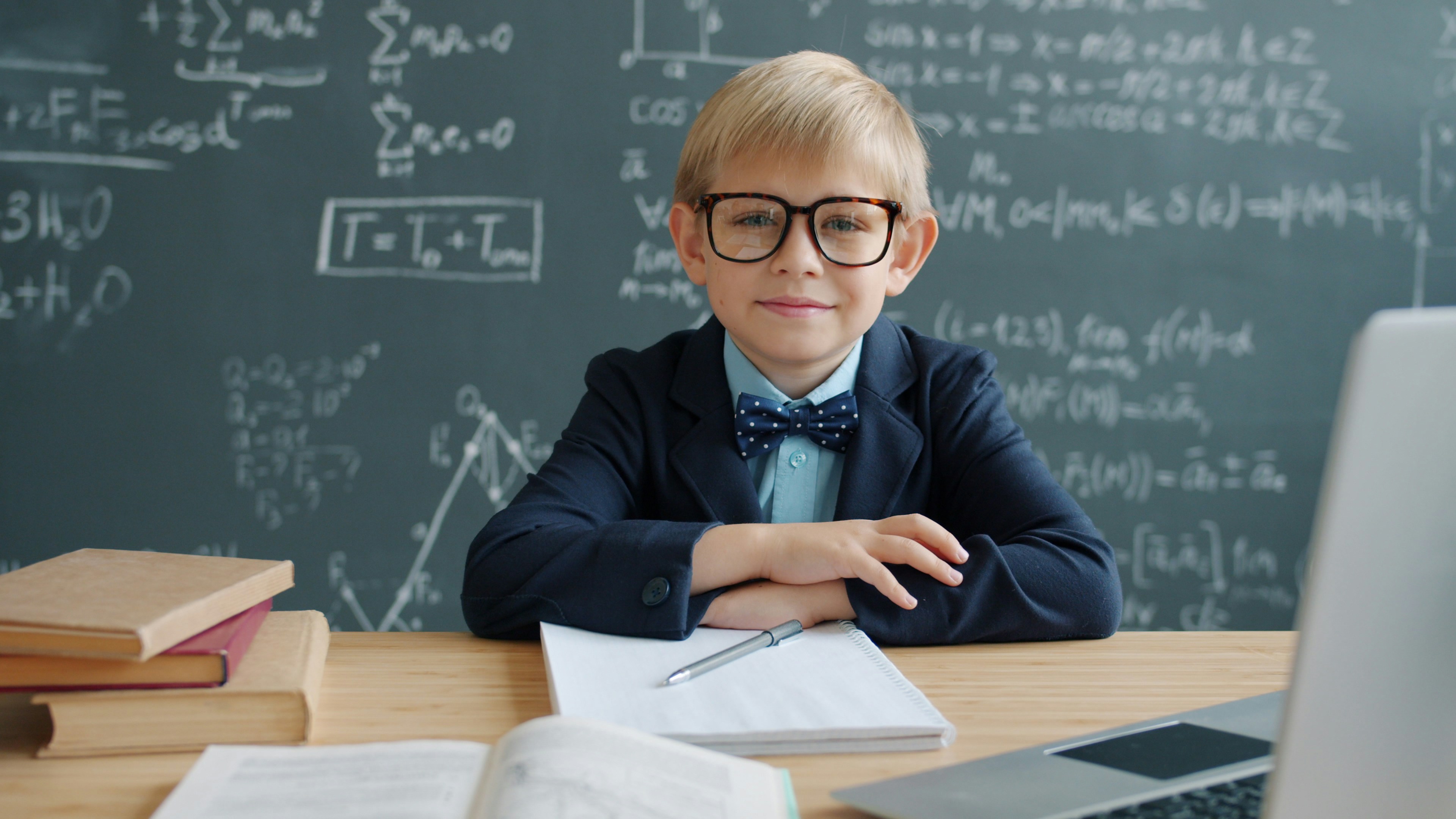 Portrait of little genius adorable boy in classroom sitting at desk wearing glasses smiling looking at camera, a chalkboard with formulae is in background.