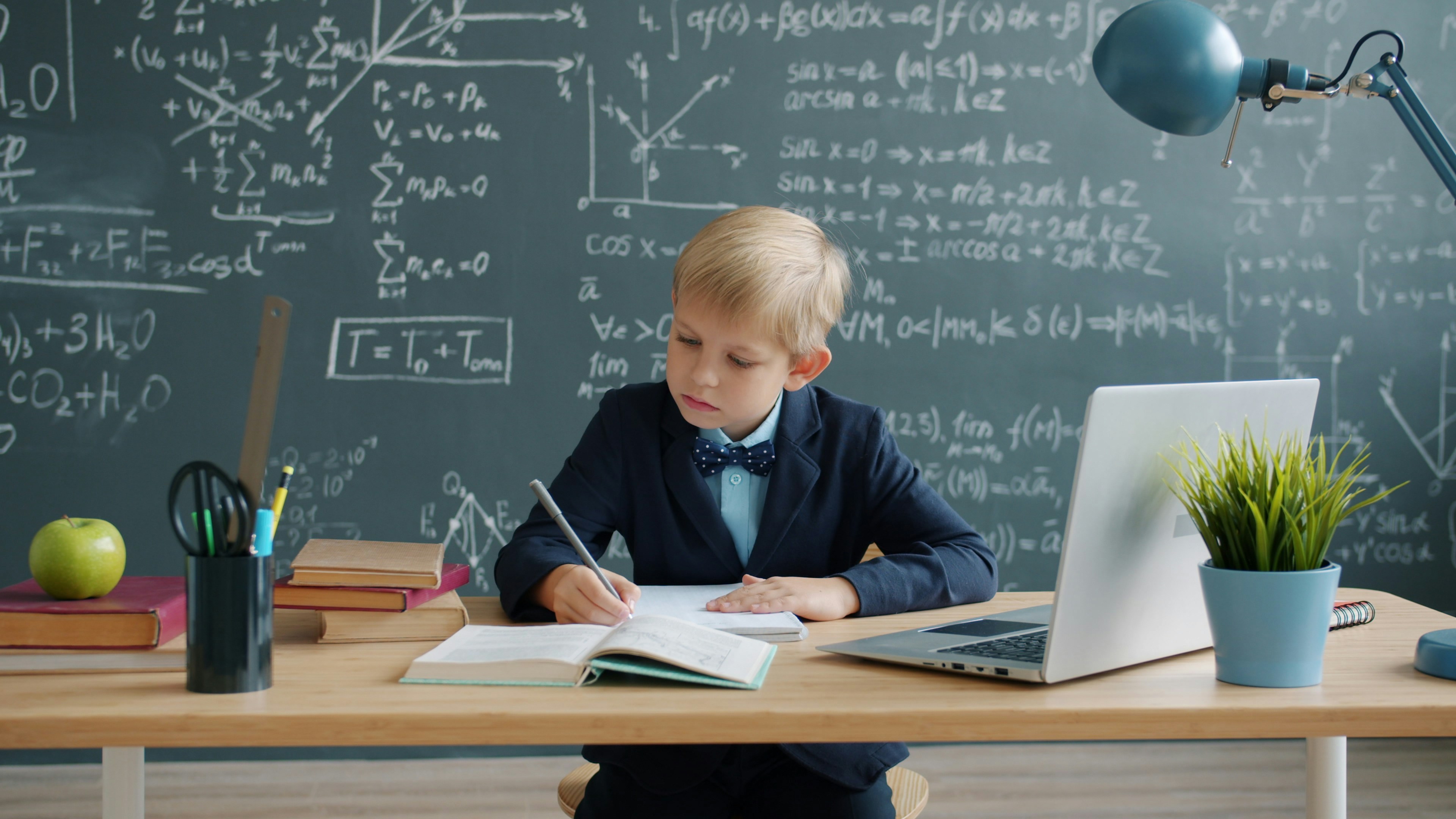 Smart little boy is writing at desk in class taking notes learning, formulas are visible on chalkboard in background. Education, prodigy kids and intellect concept.