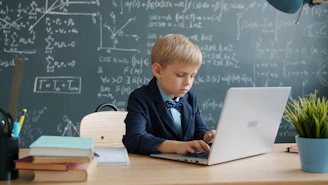Young boy in suit using laptop at desk with blackboard.