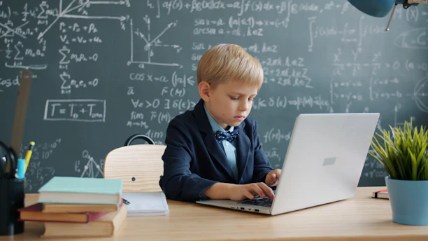 Young boy in suit using laptop at desk with blackboard.