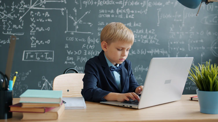 Young boy in suit using laptop at desk with blackboard.