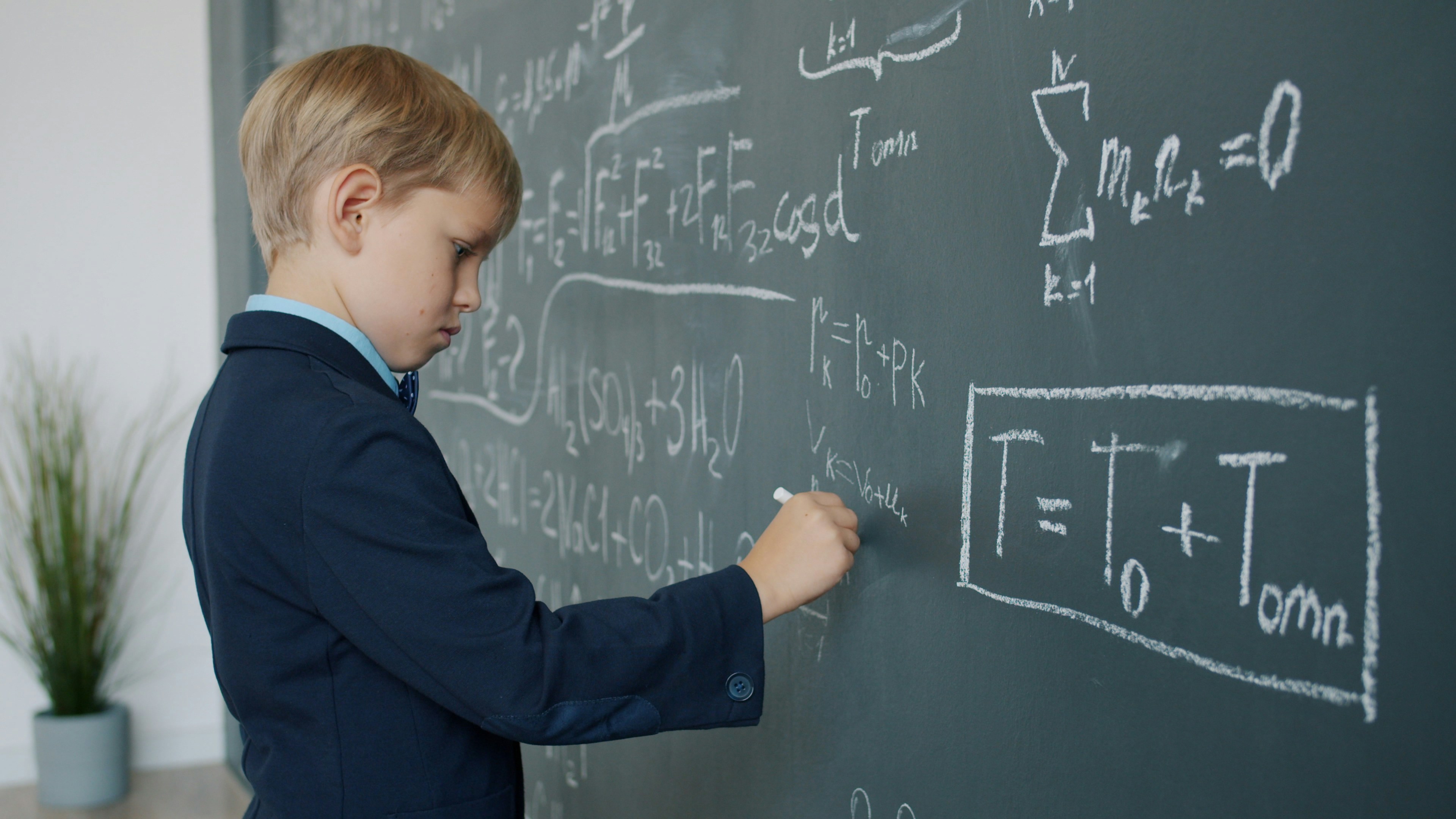 Portrait of little prodigy kid writing calculations on chalk board busy with science taking notes on blackboard learning alone in classroom. People and education concept.