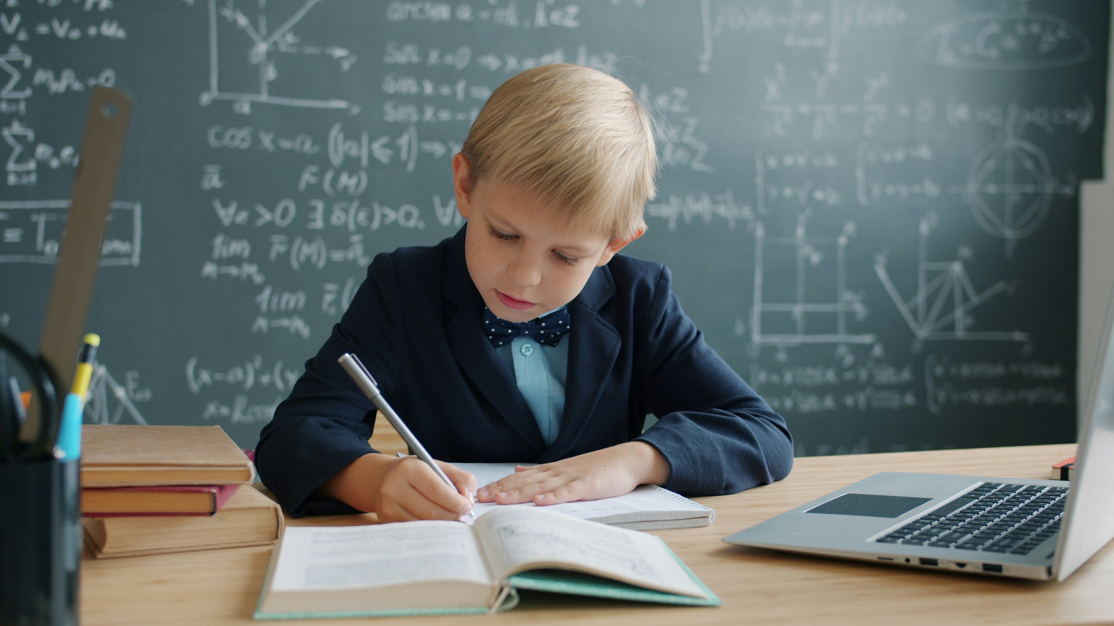 Prodigy kid in suit is taking notes sitting at desk in university classroom with chalkboard covered with formulas in background. Modern education and talanted children concept.