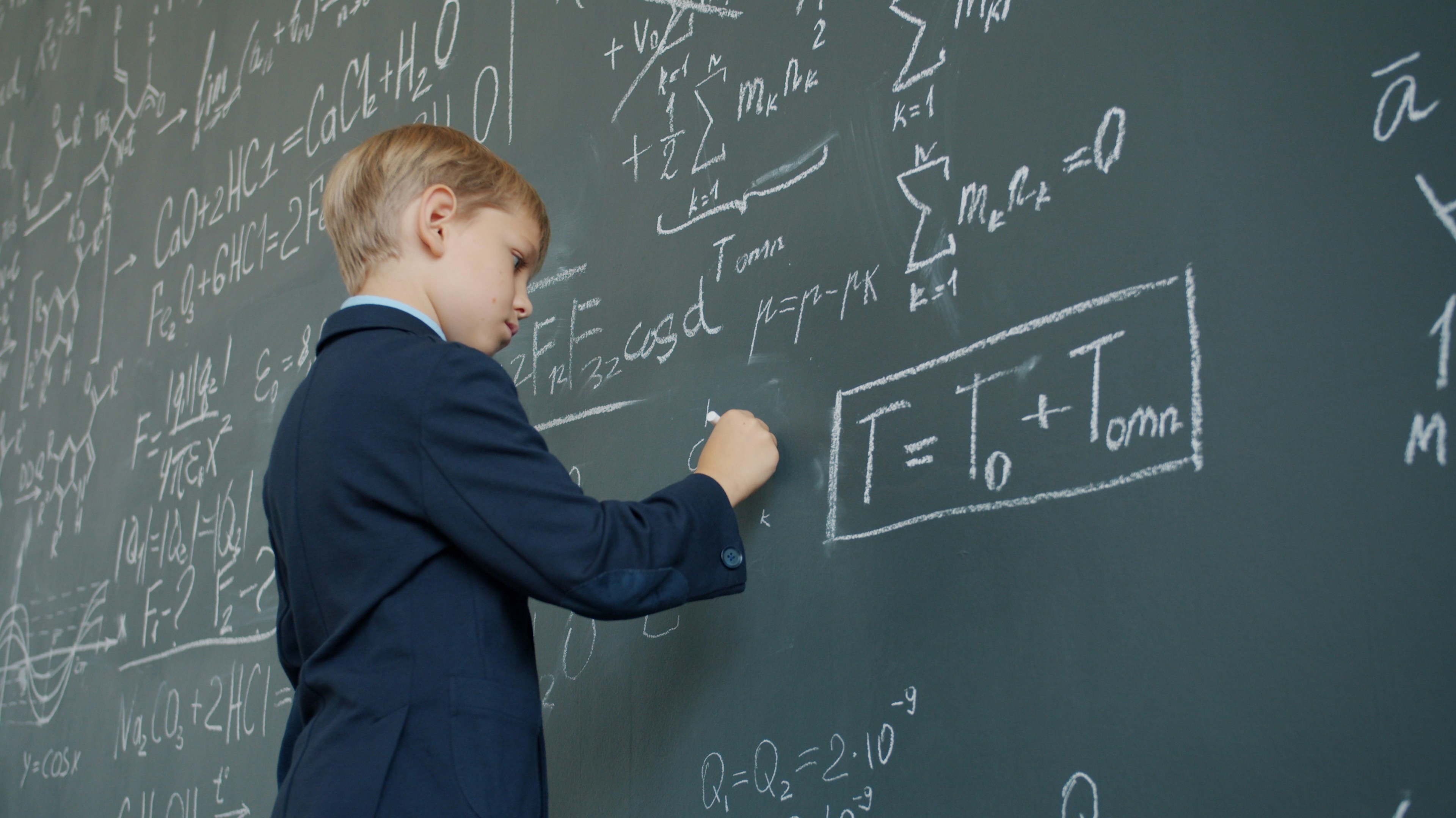 Low angle view of clever schoolboy writing formulas with chalk on blackboard focused on scientific research and education. Childhood and class concept.