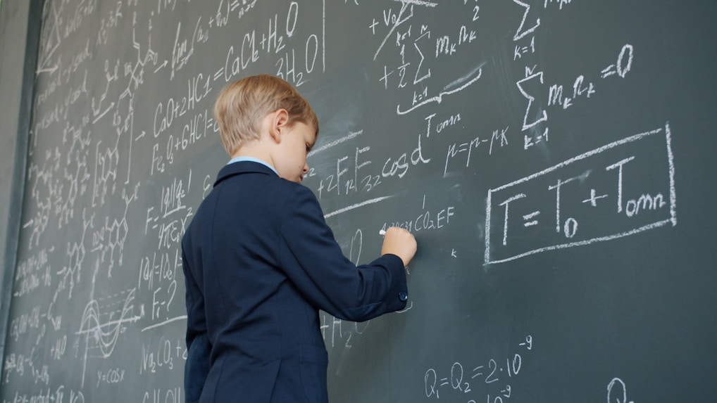 Young boy in suit writing equations on chalkboard