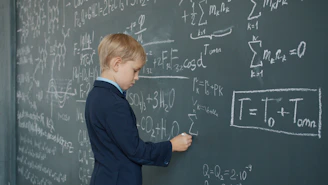 Young boy in suit writing math equations on blackboard.