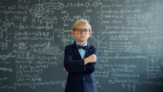Young boy in glasses stands by chalkboard with equations