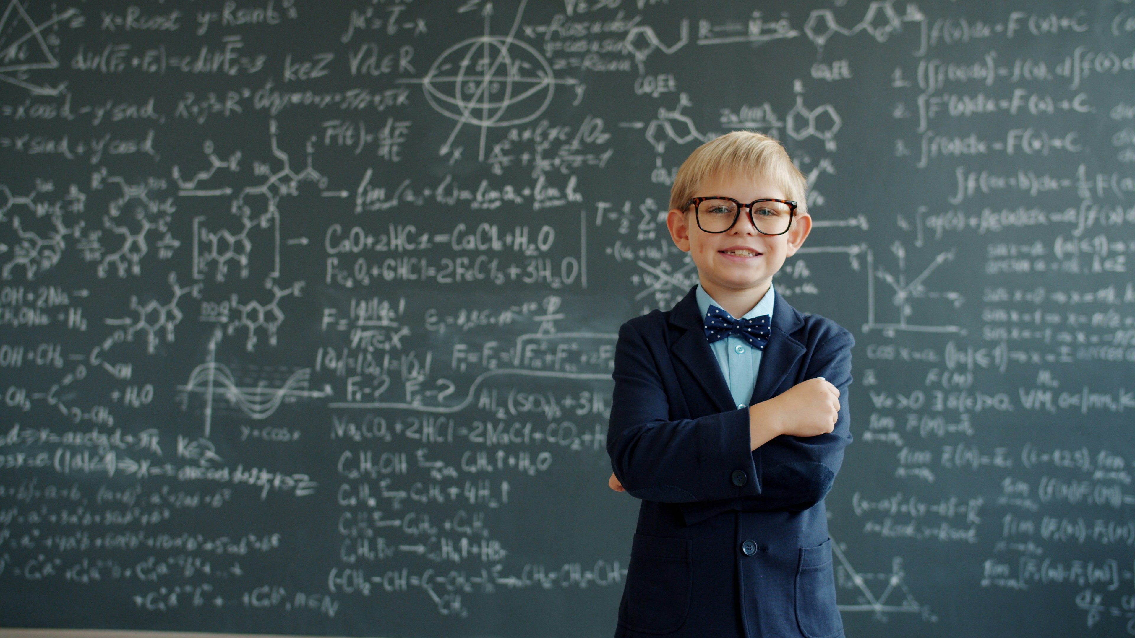 Happy kid in suit and glasses is standing in class with chalkboard in background with scientific formulas written on it. People, childhood and education concept.