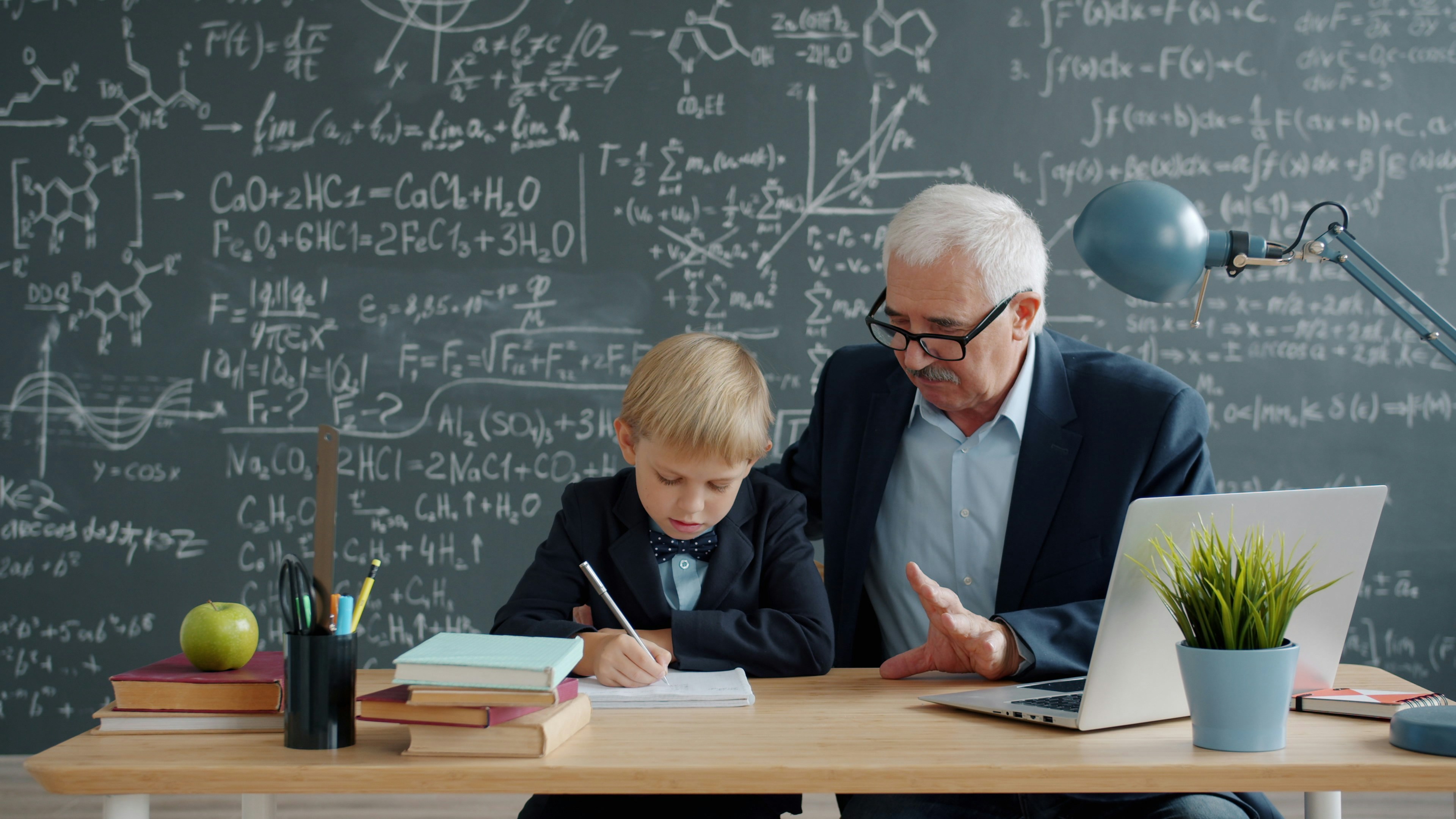 Mature man teacher in suit is teaching little smart boy in college classroom busy with education, prodigy kid is writing taking notes. People and knowledge concept.