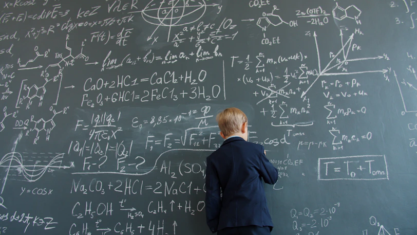 Young boy in suit writing on chalkboard with formulas