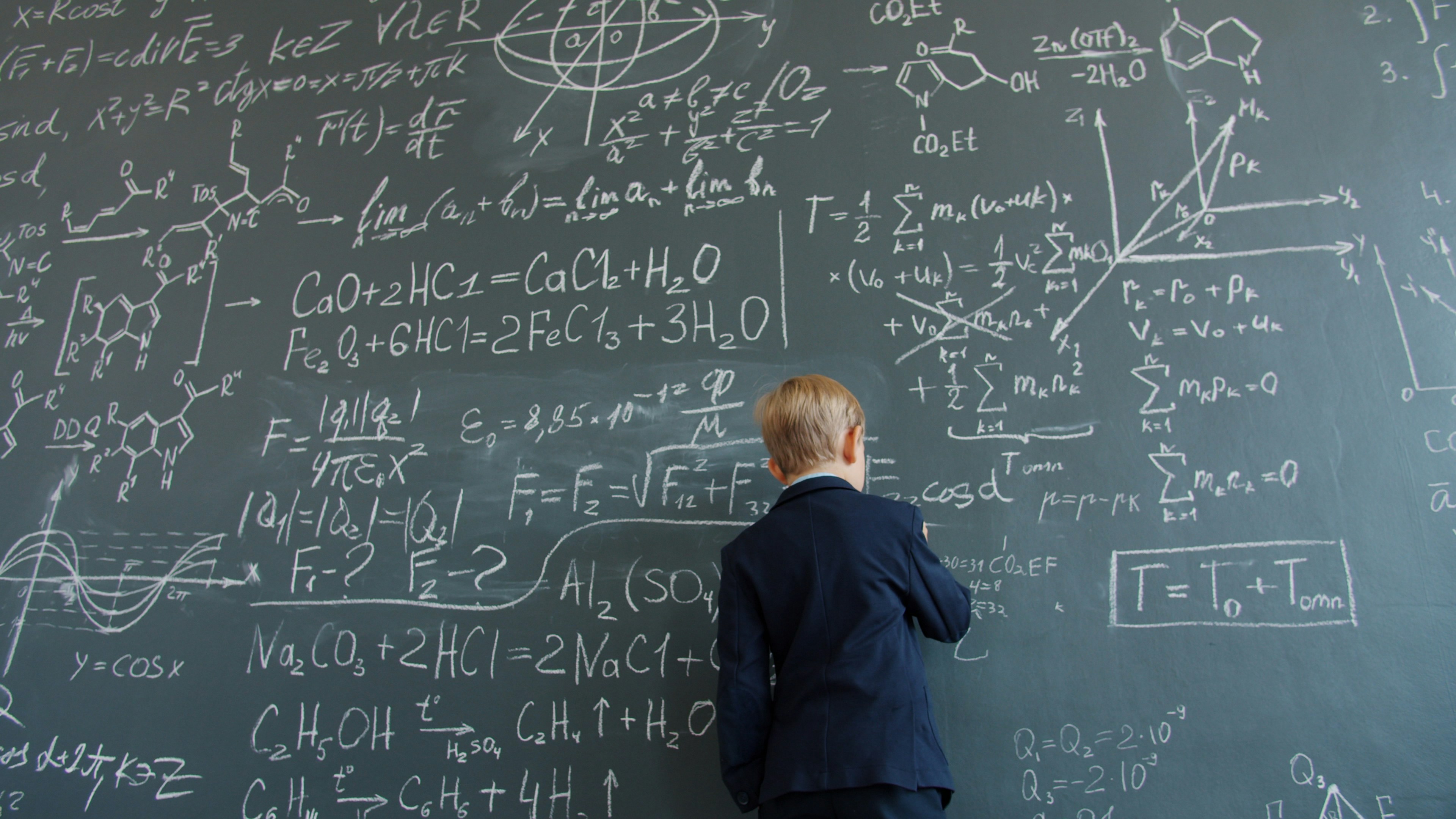 Smart little kid in suit is writing on black chalkboard busy with scientific calculations in spacious class. Modern people, education and science concept.