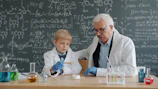 Elderly scientist teaches young boy in lab coat.