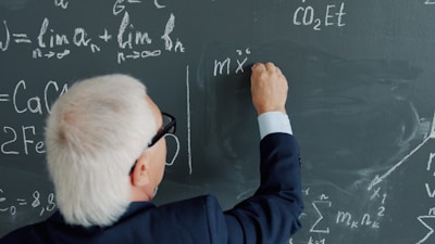 Elderly professor writing mathematical equations on a blackboard.