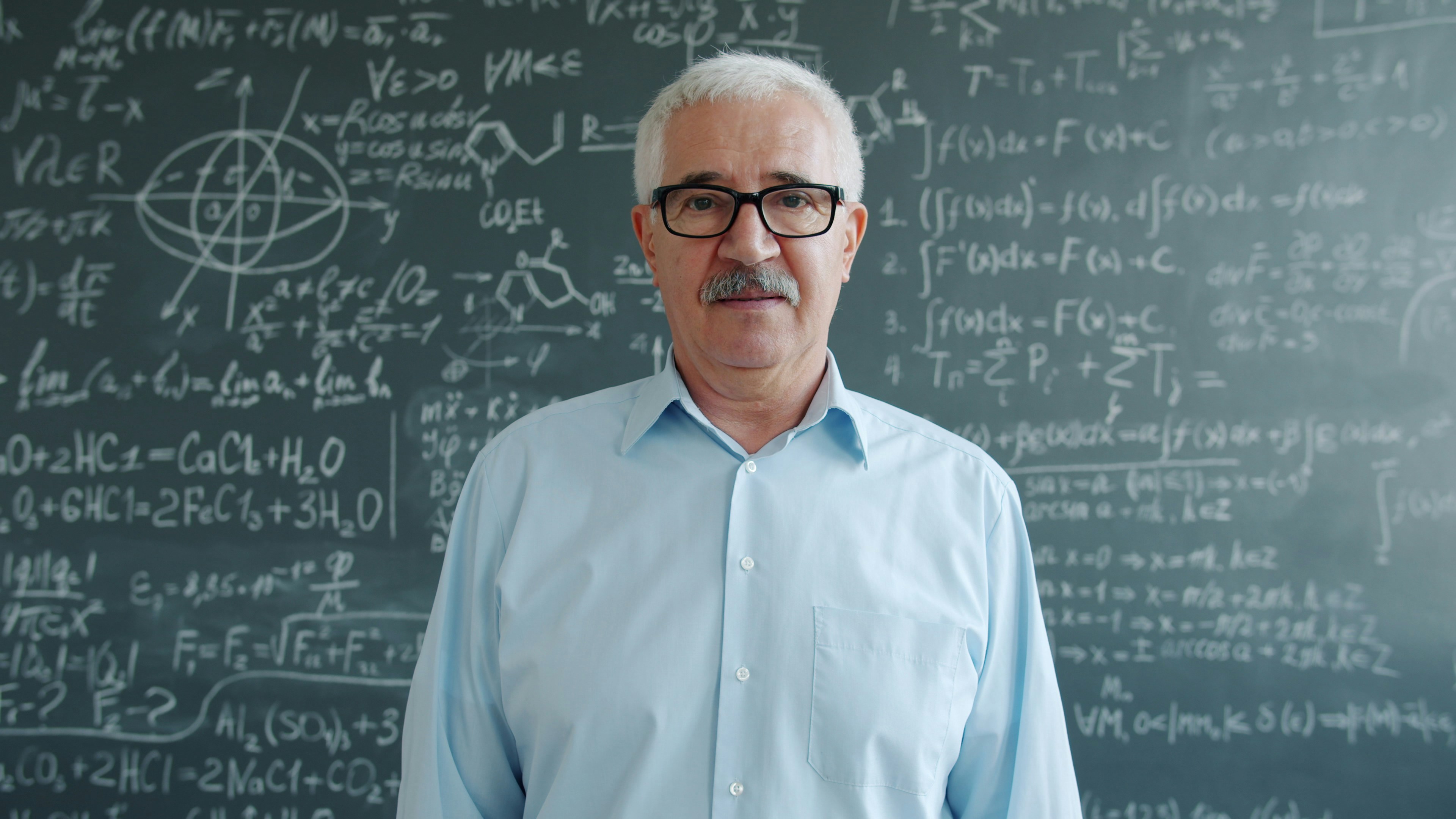 Elderly man in glasses stands before chalkboard with math equations.