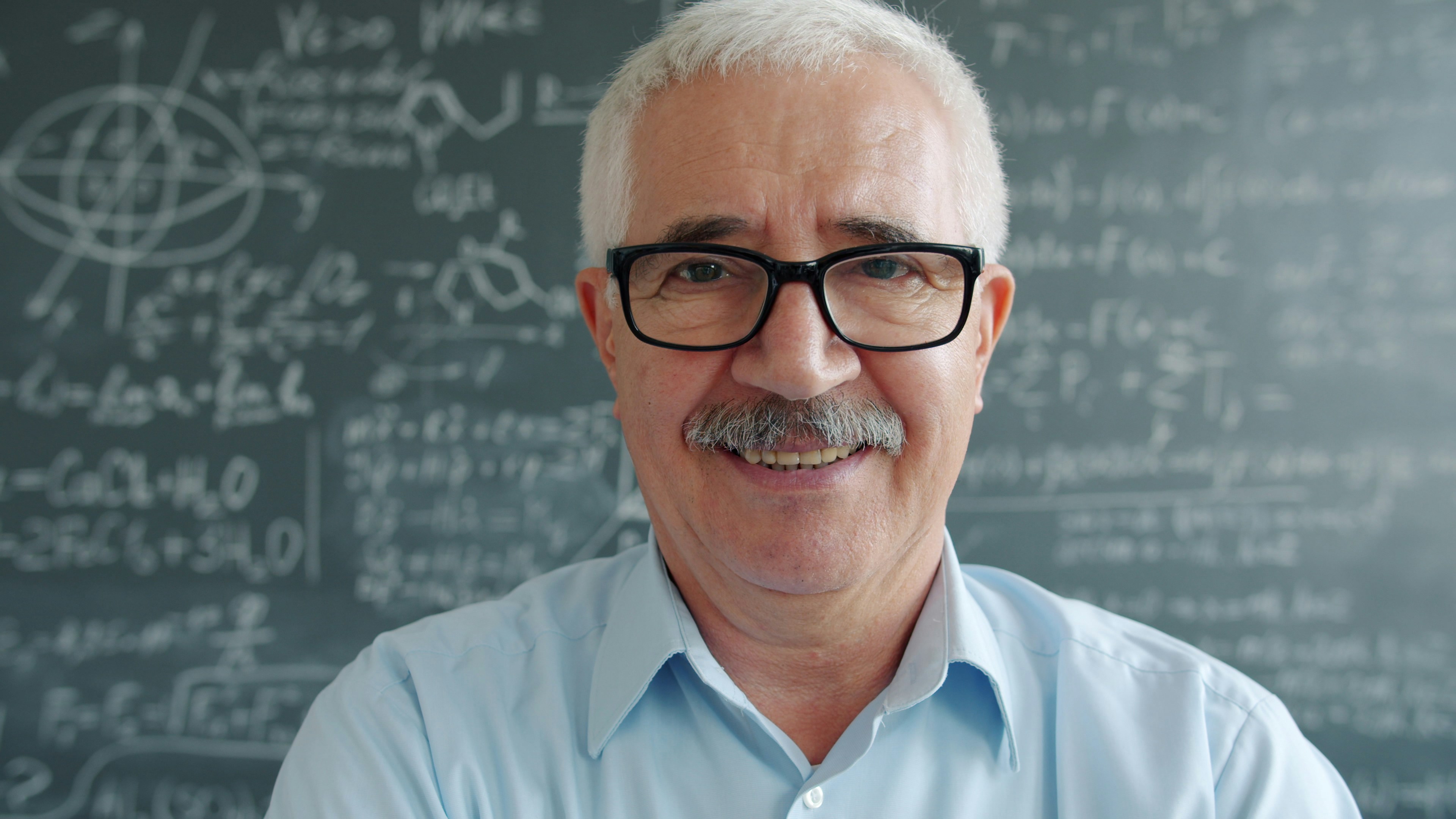 Portrait of happy mature man in glasses looking at camera smiling near chalkboard with formulas written with chalk. Education and positive emotions concept.