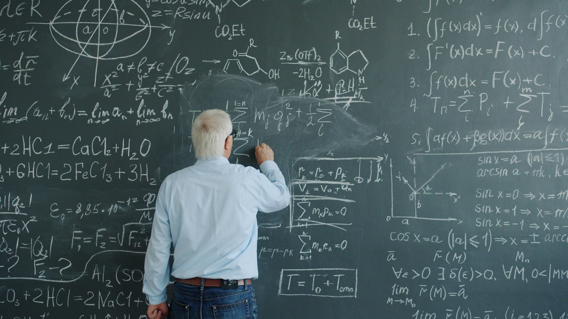 Elderly man writing complex equations on a chalkboard.