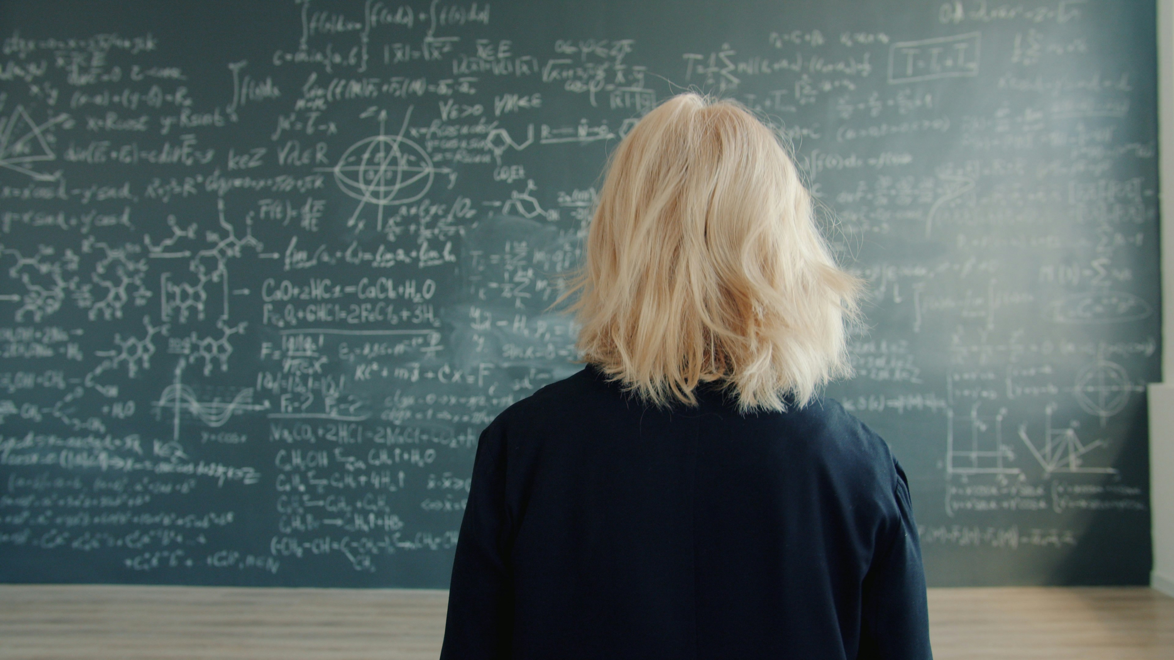 Female scientist is walking to chalkboard wall with formulas in university classroom thinking about scientific project. People and lifestyle concept.