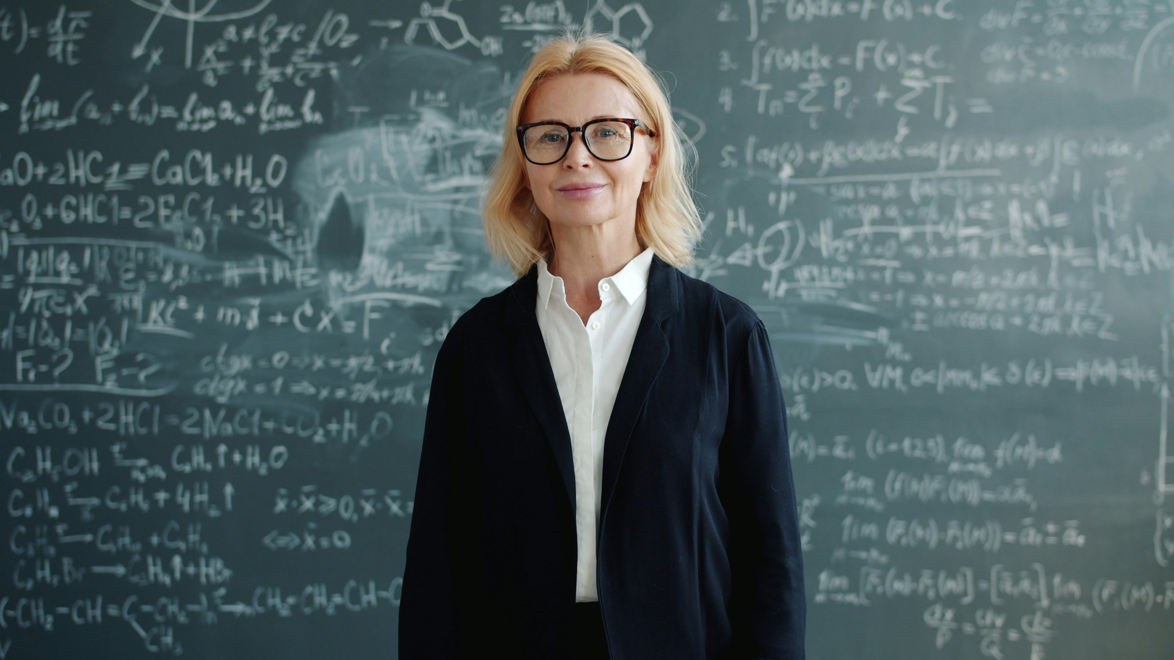 Portrait of mature adult woman wearing suit standing alone in class, smiling and looking at camera, blackboard with formulas is visible in background