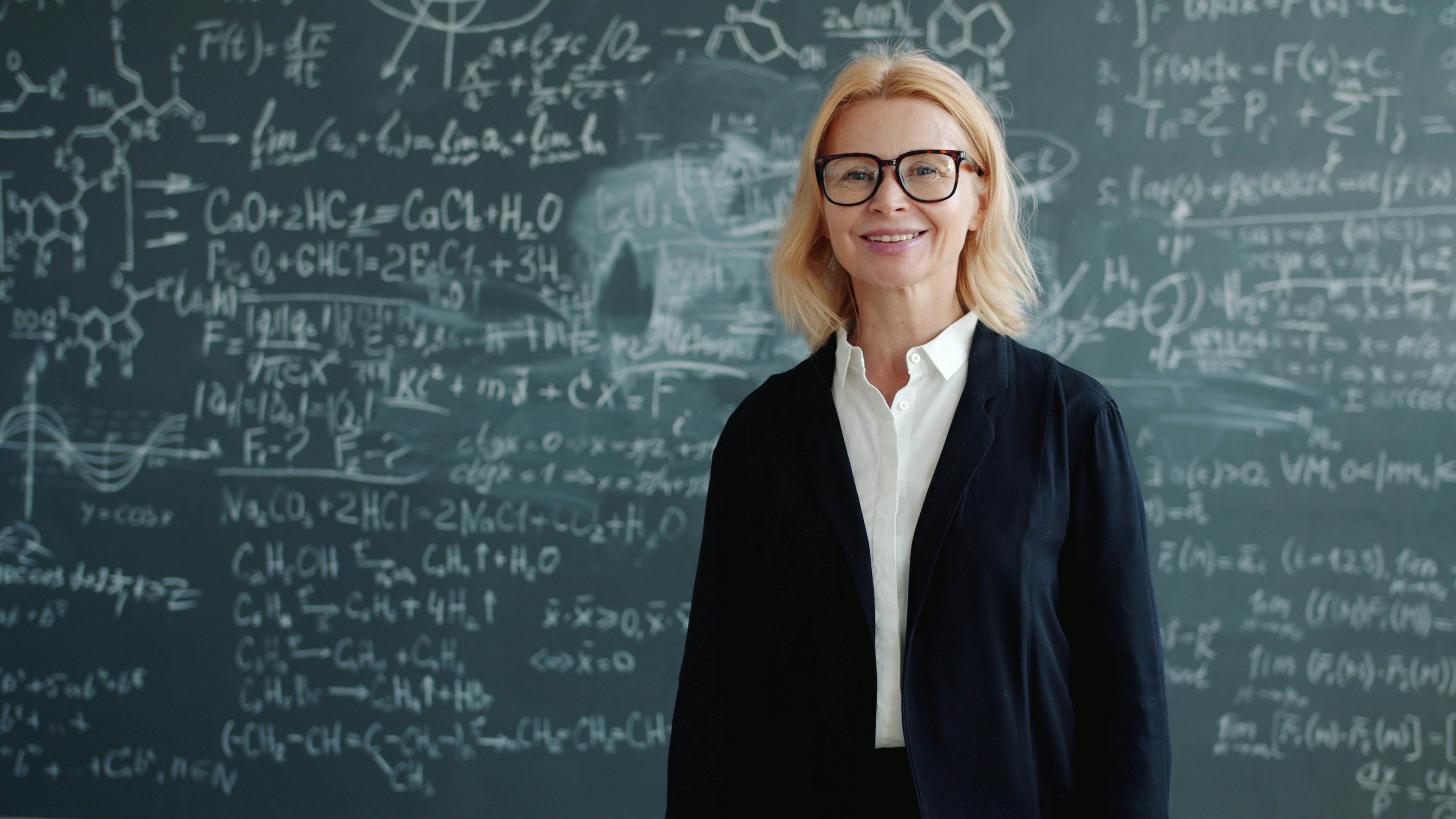 friendly teacher wearing suit and glasses smiling looking at camera standing alone in classroom, blackboard with scientific formulas is in background.