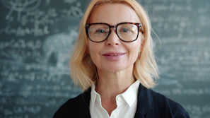 A woman with glasses in front of a chalkboard.