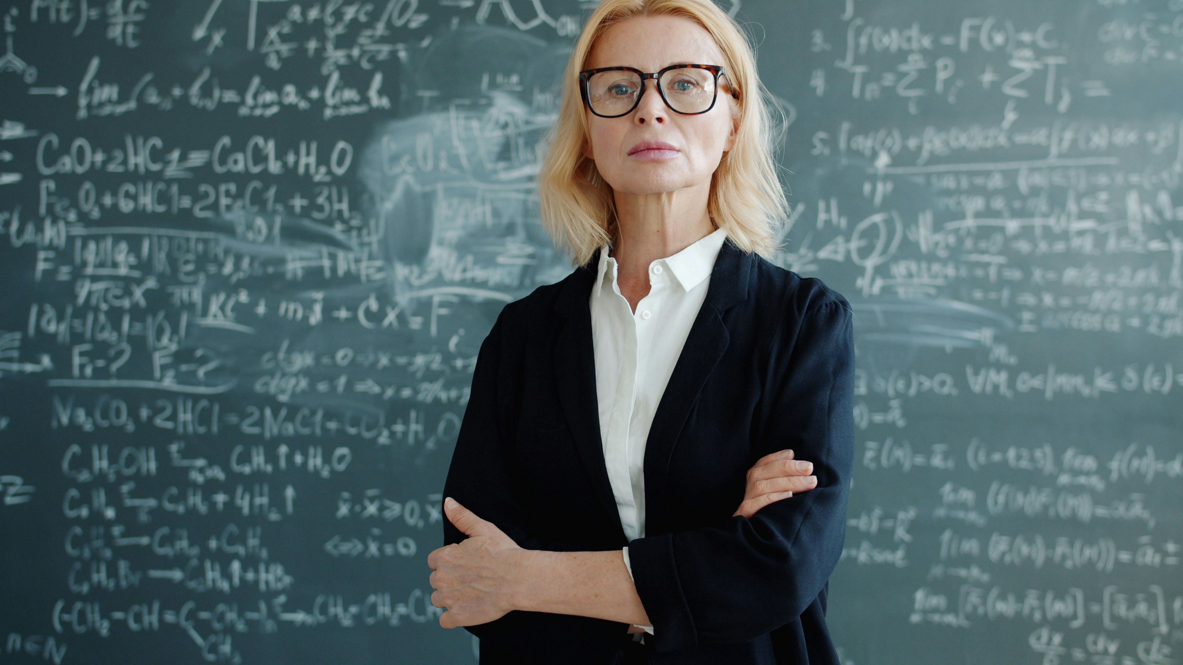 Portrait of serious smart lady professor in classroom with formulas on chalkboard standing alone with arms crossed looking at camera. People and education concept.