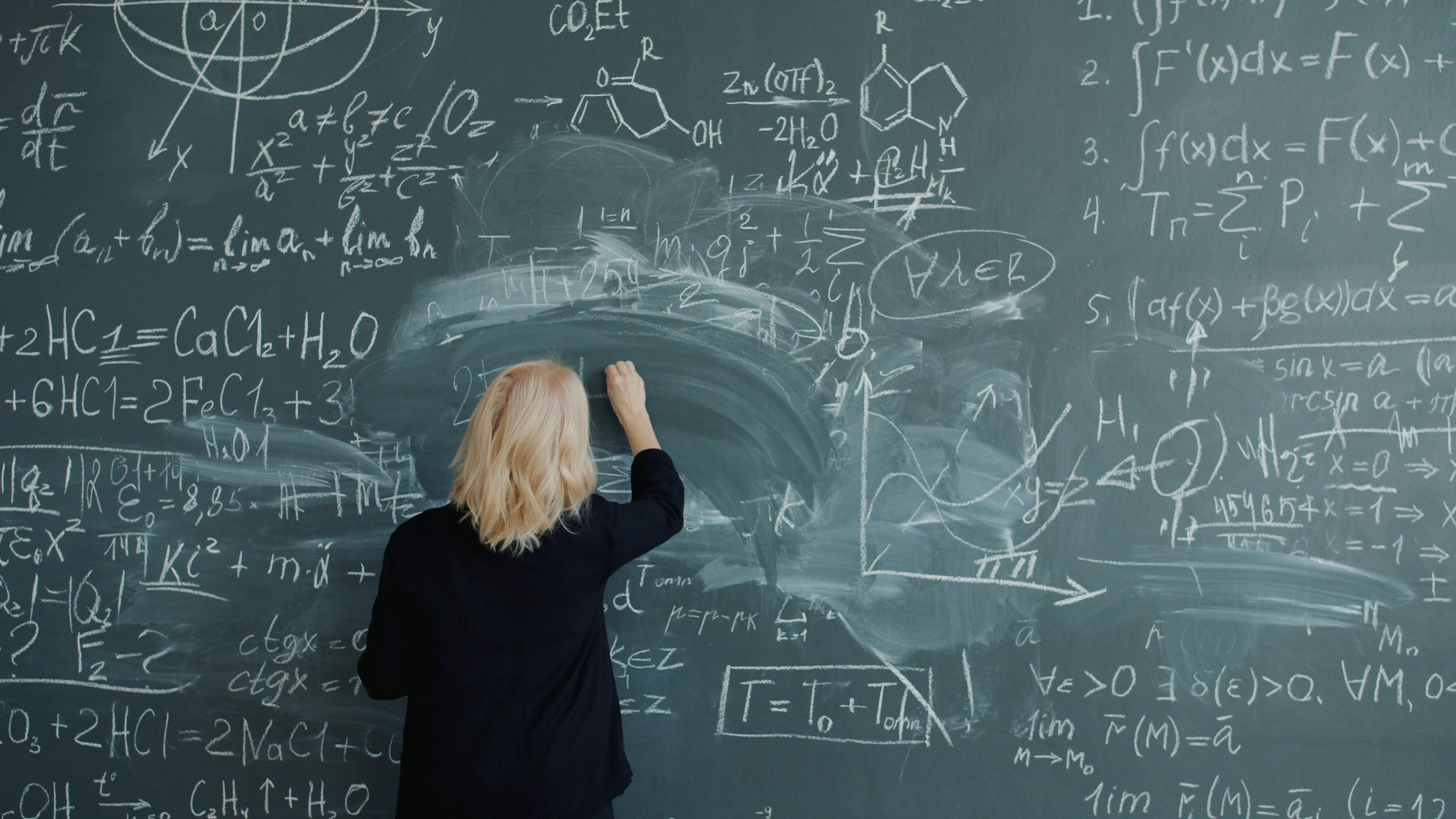 Adult woman teacher in elegant suit and glasses is writing formulas on blackboard talking teaching class at school. Education and occupation concept.