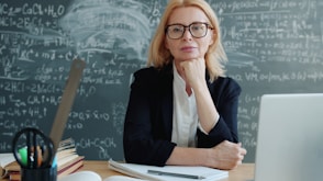 Teacher sitting at a desk with chalkboard formulas
