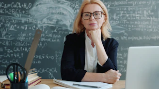 Teacher sitting at a desk with chalkboard formulas