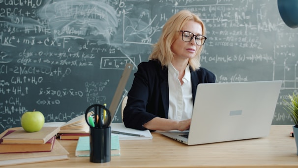 Teacher working on laptop in front of chalkboard.