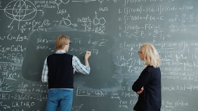 Student writes math equations on blackboard while teacher watches.