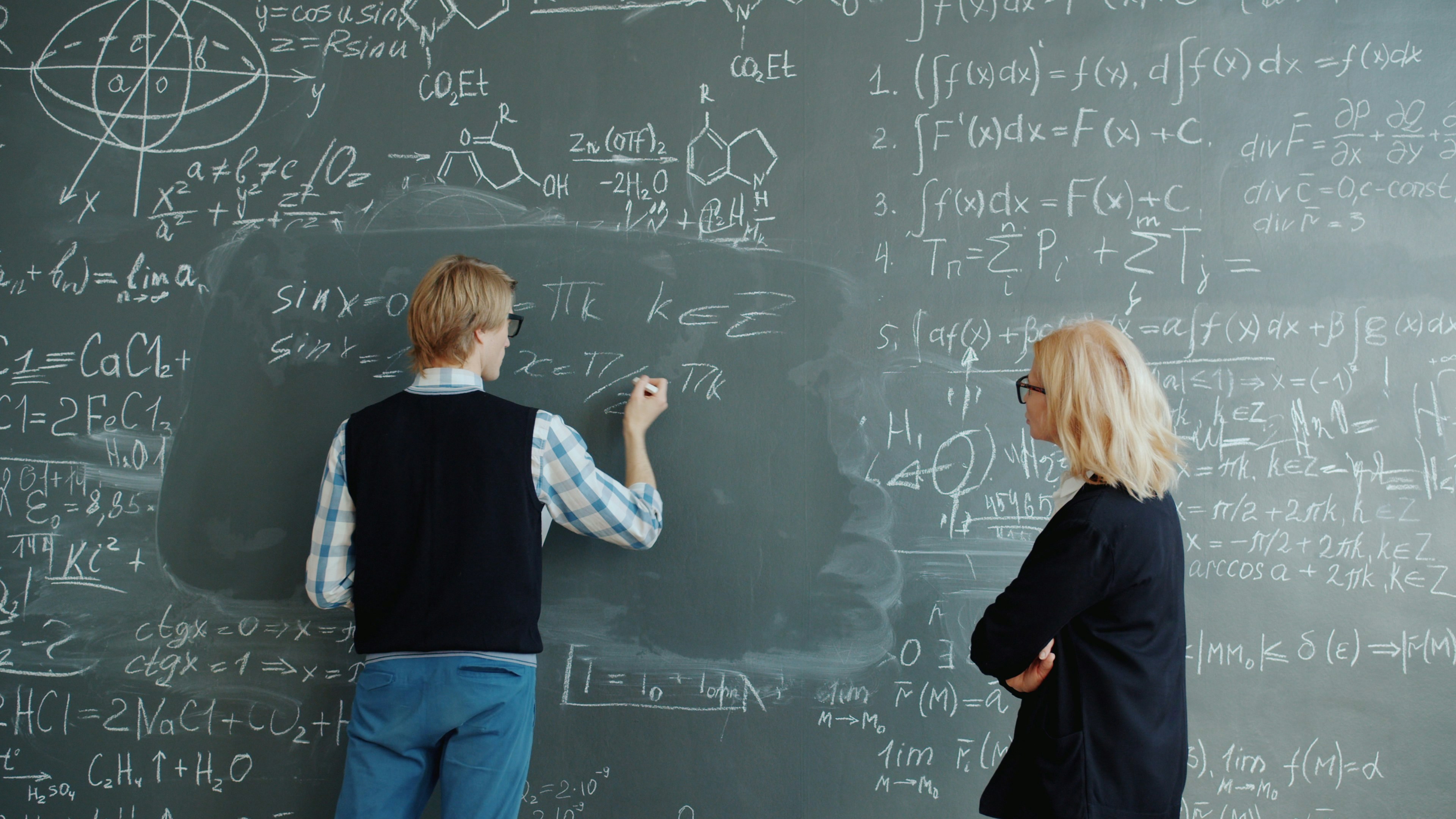 Student and helpful female teacher are working with chalkboard writing formulas in classroom, woman is teaching the guy talking explaining information.