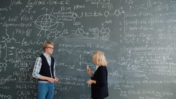 Two people discussing math formulas on a blackboard.