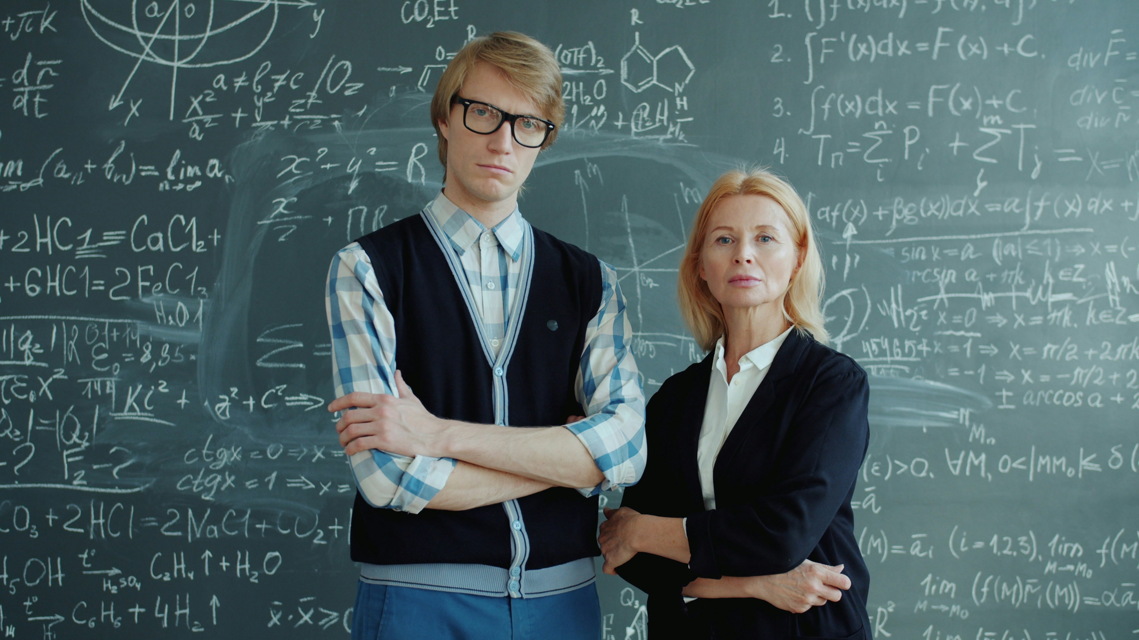 Portrait of man and woman standing near chalkboard with formulas looking at camera with serious faces holding arms crossed. Education and people concept.