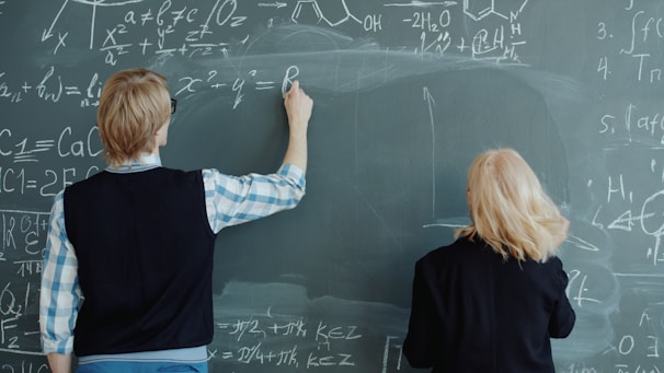 Two people writing math formulas on a chalkboard.