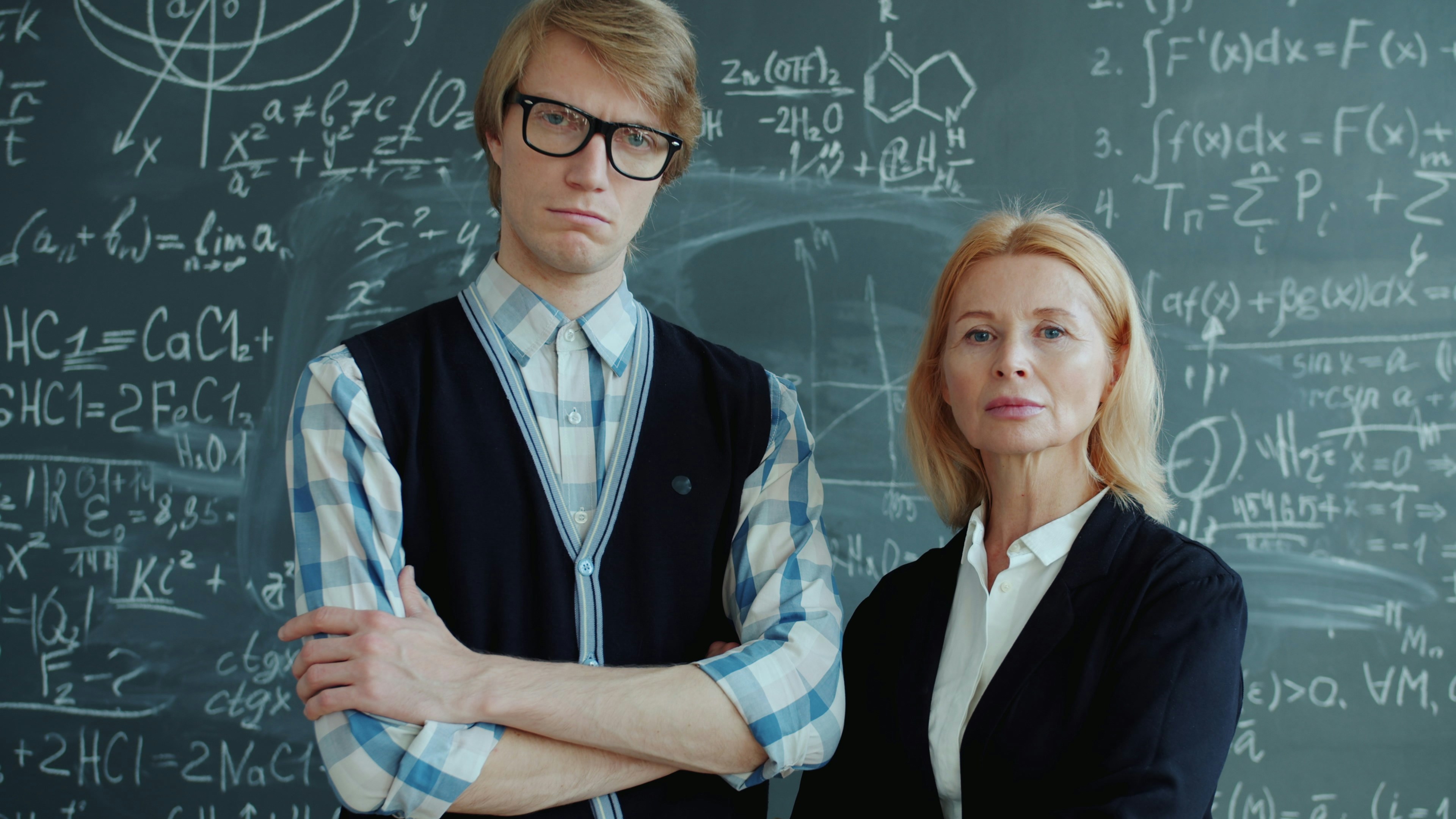serious teachers male and female looking at camera indoors standing with arms crossed with chalkboard in background. People and lifestyle concept.