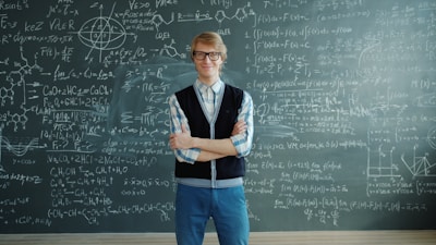 Man in glasses stands before a chalkboard with equations.