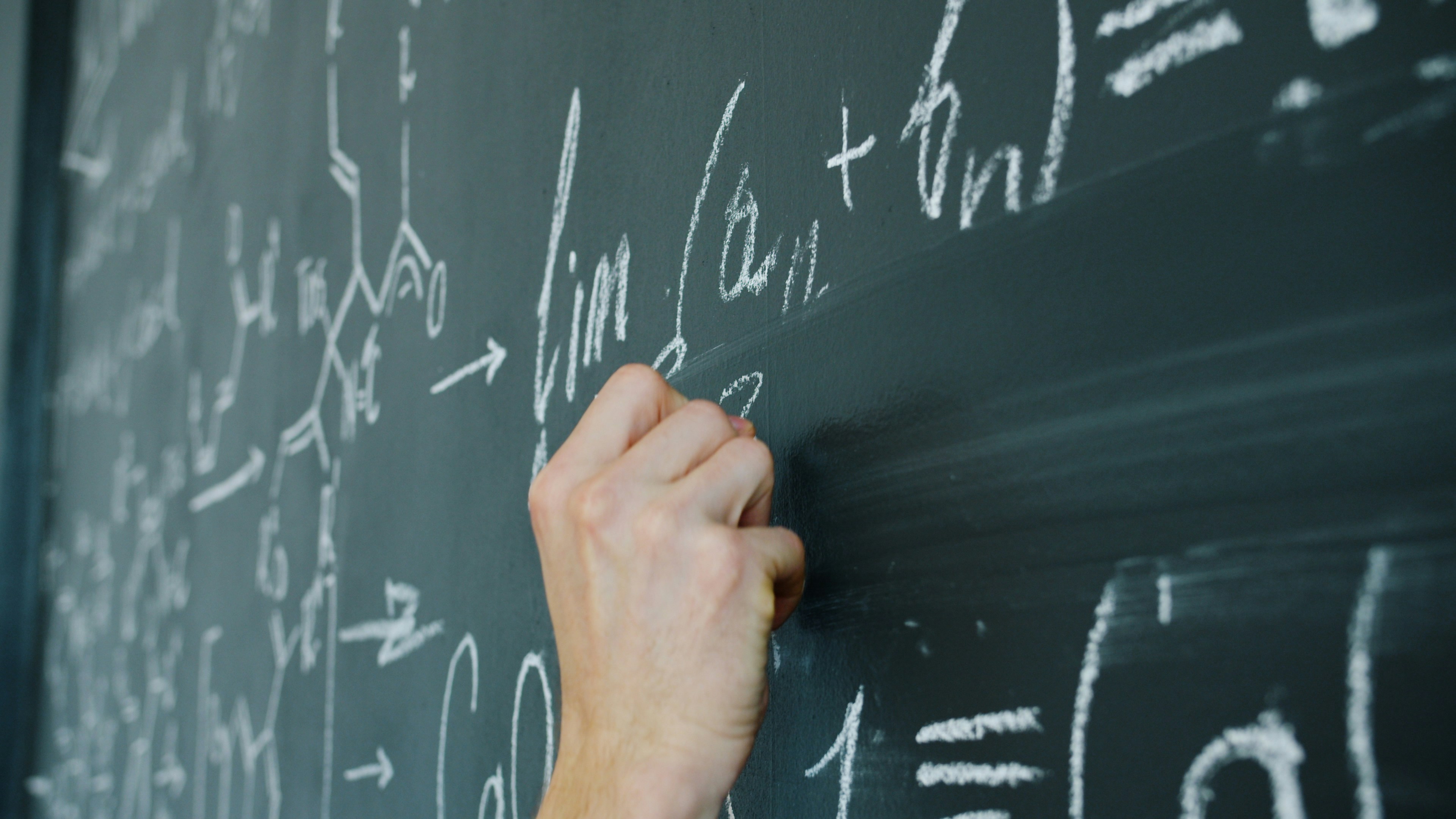 maths student's hand writing formulas with chalk on blackboard indoors in classroom. Education, people and modern science concept.