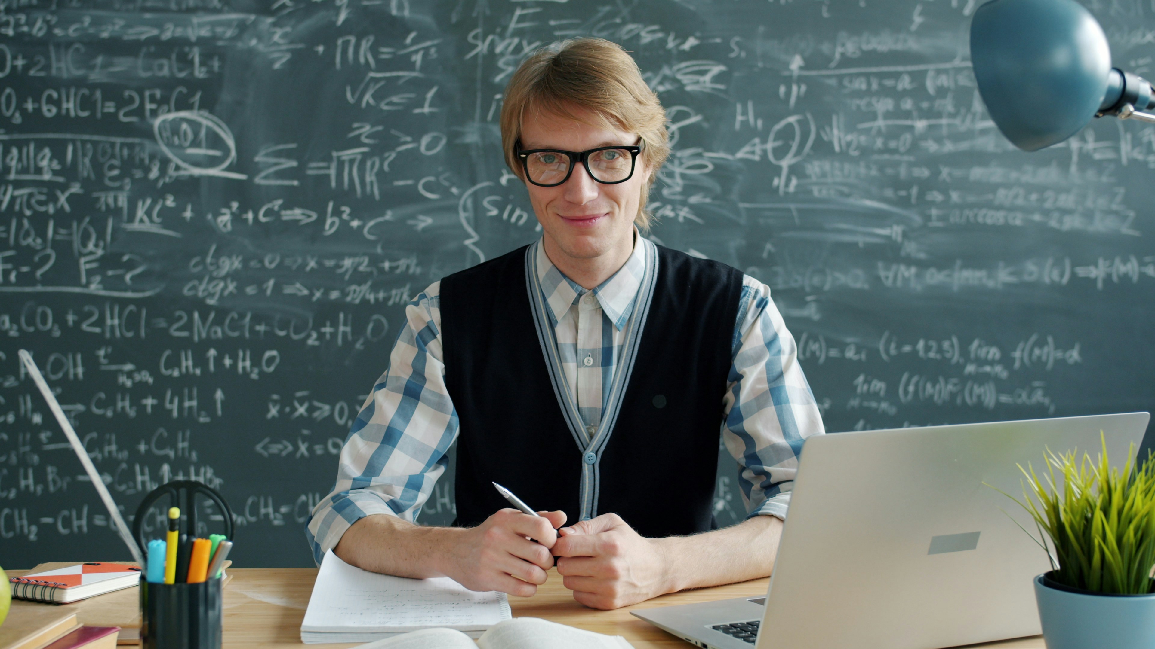 Portrait of friendly young man teacher smiling sitting at desk in class looking at camera, laptop and books are visible on table. People and education concept.