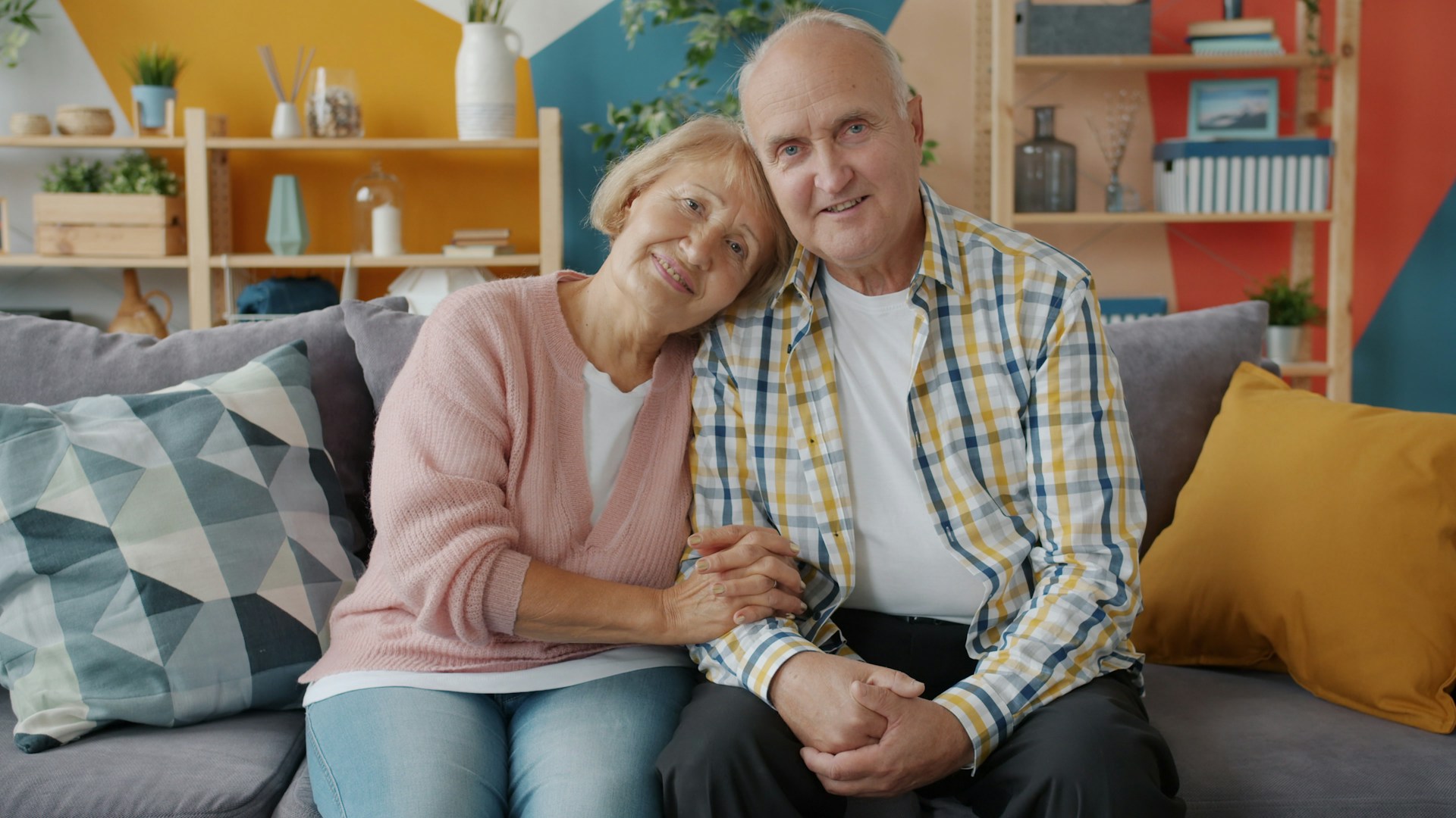 Elderly couple smiling and holding hands on couch.
