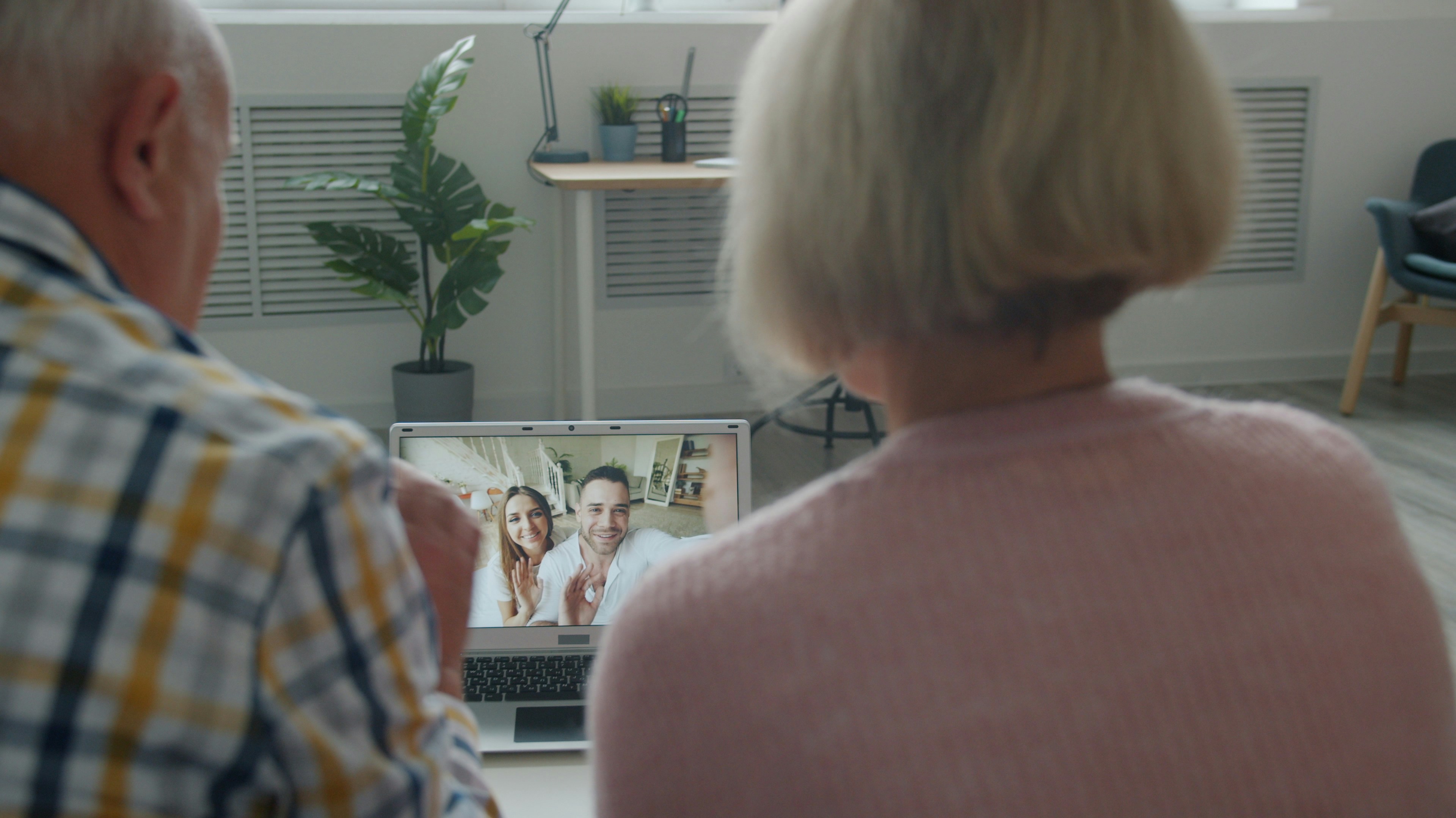 Back view of senior couple making online video call with laptop from home talking to young people using modern device. Family and communication concept.