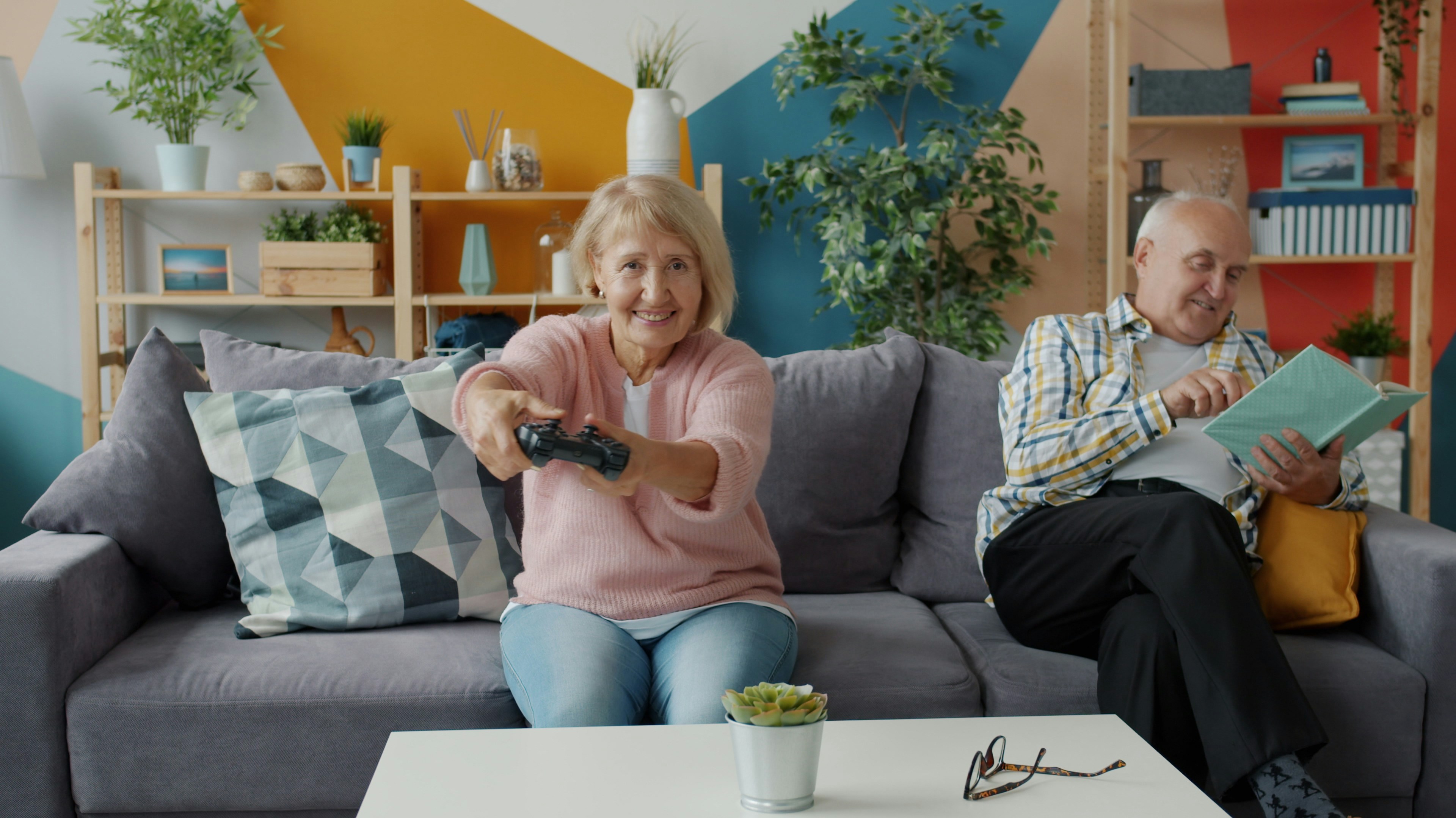 Portrait of senior woman playing video game having fun while old man is reading book relaxing on couch at home. People, relaxation and hobby concept.
