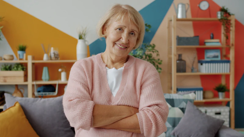Smiling elderly woman with arms crossed in living room.