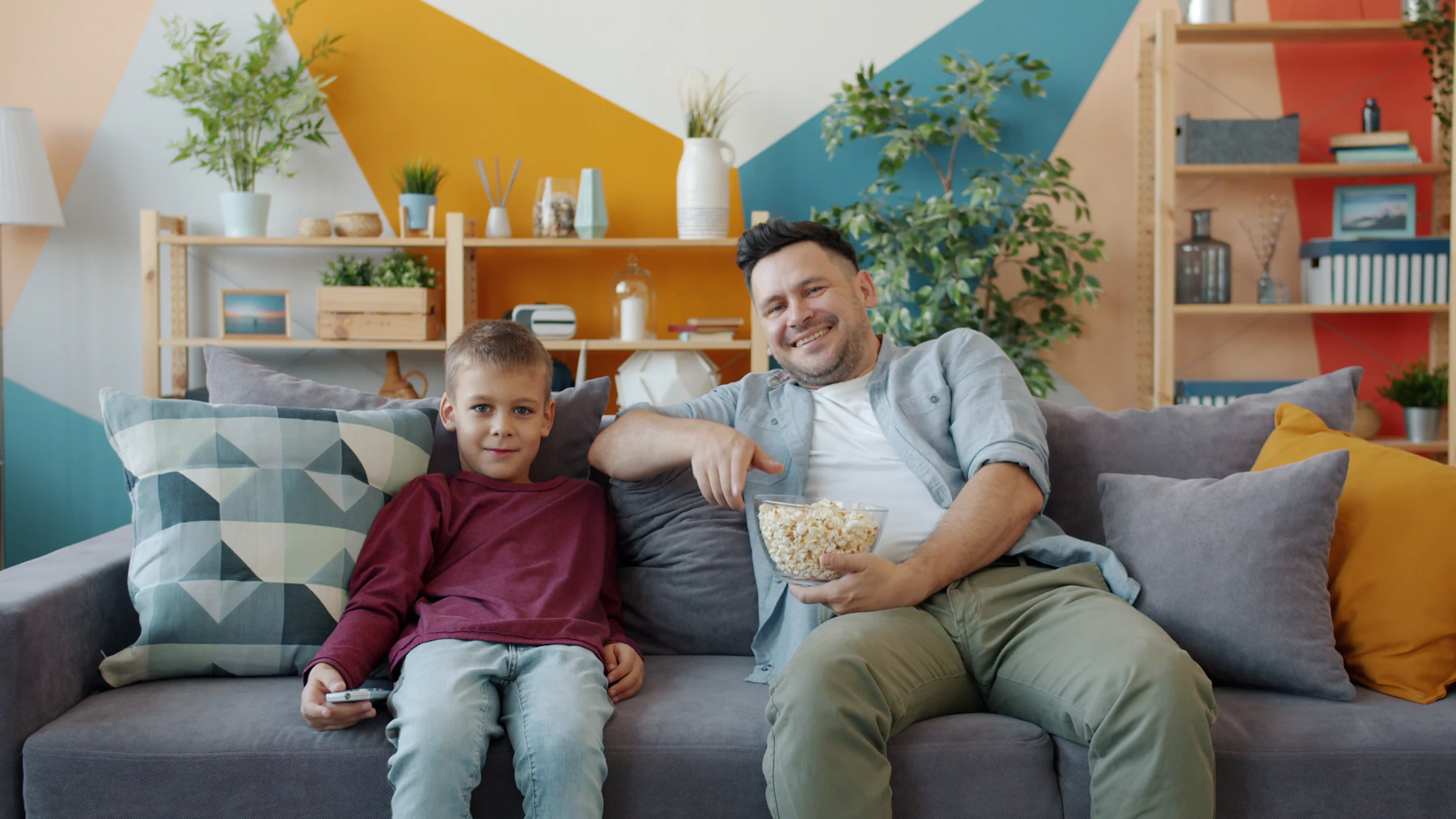 A father and son on a sofa watching TV with a bowl of popcorn