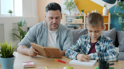 Father reading book while son writes at table.