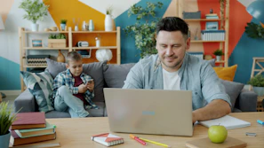 Father works on laptop while son plays on phone.