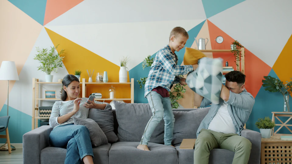 A boy jumping on a living room sofa in a playful pillow fight with his father while his mother sits beside them